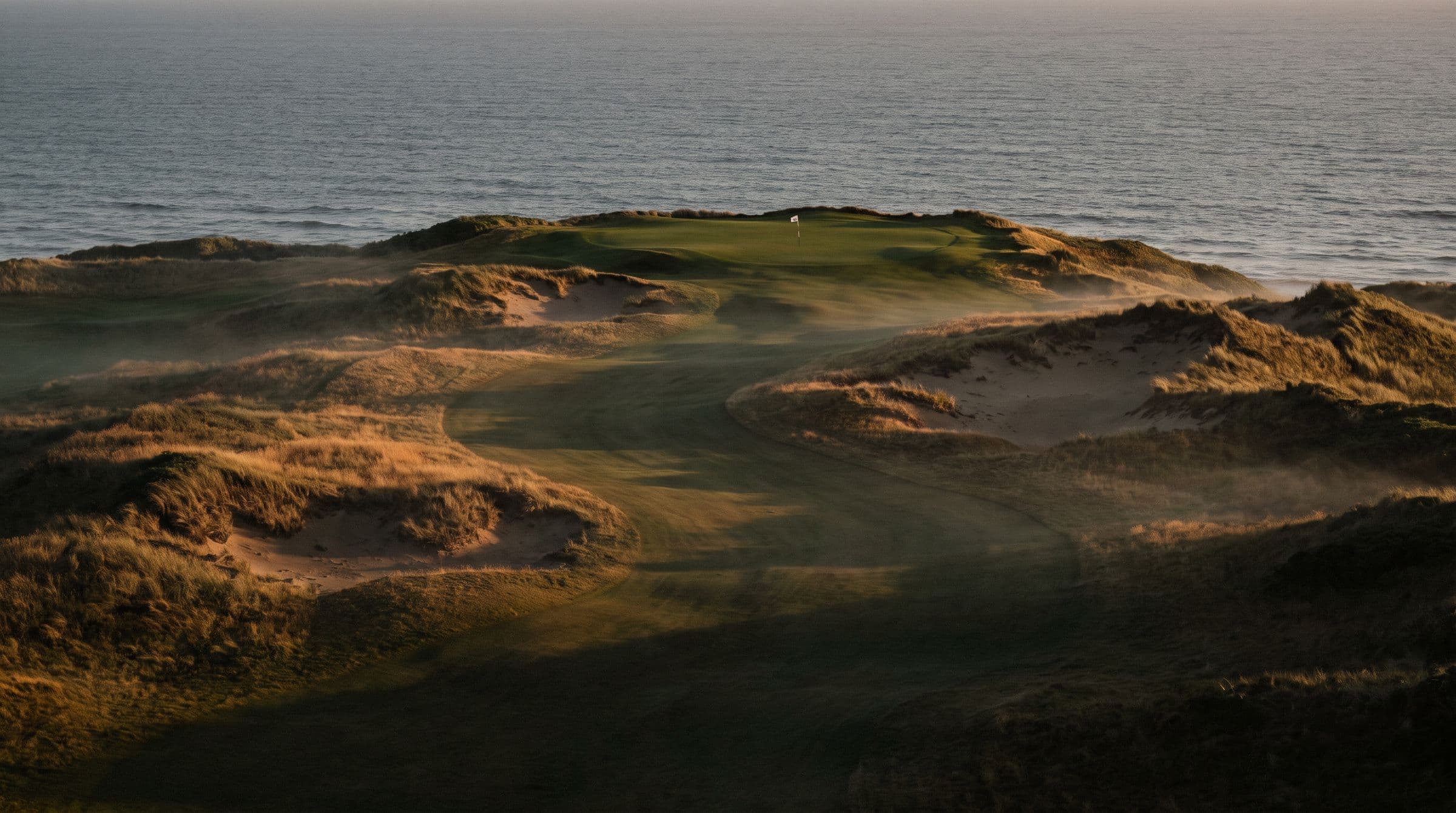 Dusk light over a links fairway winding between dunes toward a green on the headland, the sea beyond.