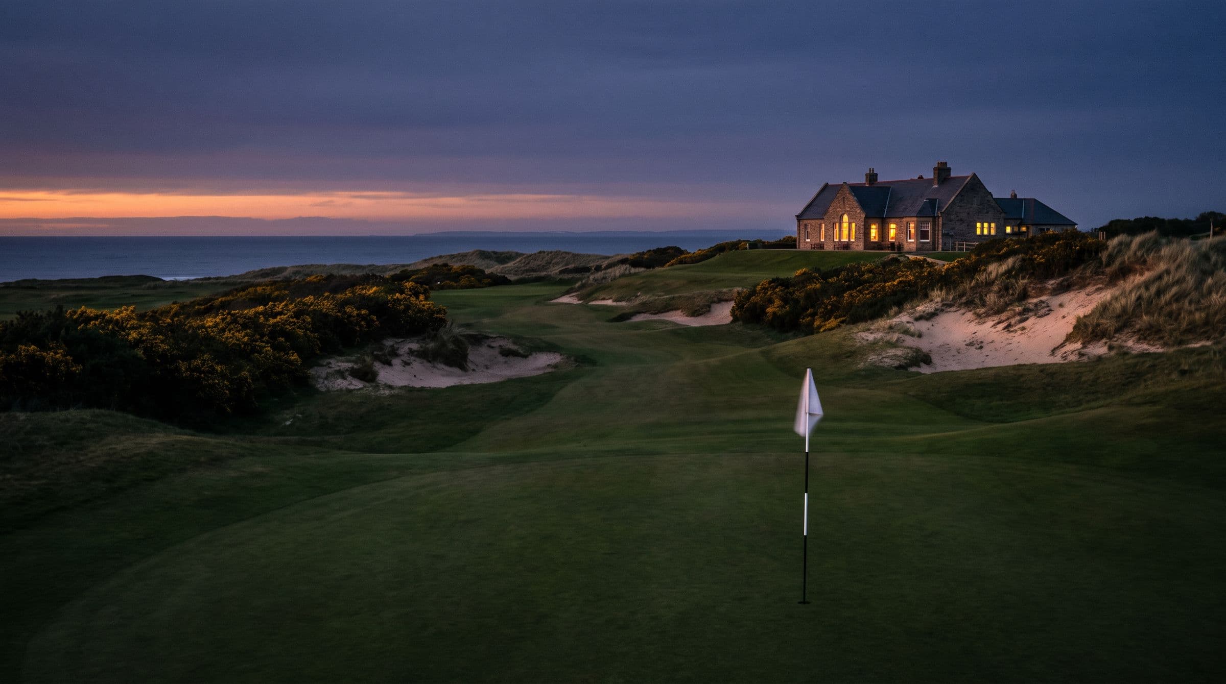 The final fairway rising to a lit clubhouse in the last orange light.