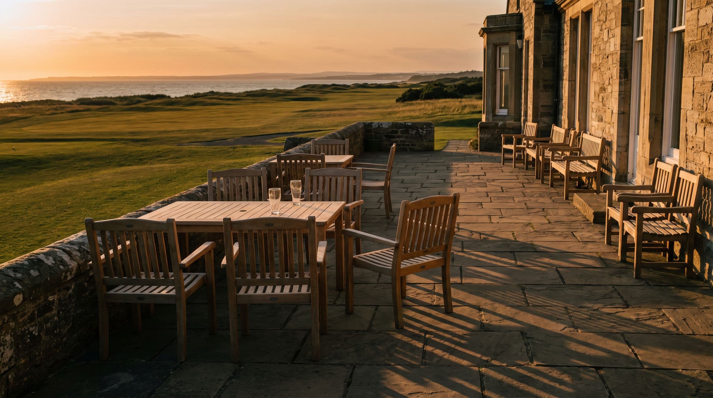 An empty stone terrace with teak tables in golden light above the fairway and sea.